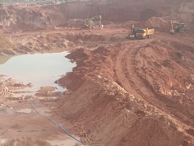 The image depicts an active construction site, likely a quarry or large excavation project, in Kwang, Nigeria. The terrain is characterized by large mounds of reddish-brown earth and exposed rock faces, indicating significant earthmoving operations. In the foreground, a body of murky water has accumulated, reflecting the overcast sky. Several pieces of heavy machinery are visible, including excavators and a large dump truck, suggesting ongoing excavation and material transport. A blue hose runs along the ground, possibly for water management. The background shows more layered earth and distant structures, hinting at the scale of the development. The scene is devoid of people, emphasizing the mechanical nature of the work.