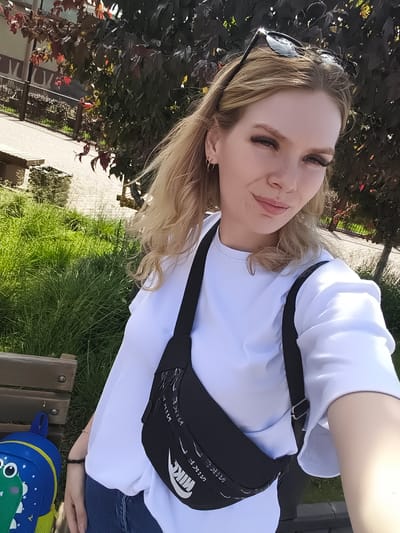 A young fair-skinned woman, appearing to be in her mid-20s to early 30s, is centered in a selfie taken outdoors in bright daylight. She has long blonde hair, styled away from her face, with dark sunglasses perched on her head. She wears small gold-colored hoop earrings. Her eyelashes are long and dark, and she exhibits a slight, closed-mouth smile while looking directly at the camera. She is dressed in a white long-sleeved crew-neck shirt and blue denim jeans, partially visible on her lower left. A black fanny pack, prominently featuring a white Nike swoosh logo and repeating "NIKE" text, is worn crossbody over her right shoulder and rests on her torso. A thin, dark bracelet is visible on her left wrist. To her left, on a wooden slatted bench, a blue backpack with a green cartoon dinosaur character is resting. The immediate ground is paved with light-colored stones, transitioning to dense green grass on the left. In the background, behind a dark metal fence, are trees with dark green and some reddish foliage. A building or structure is partially visible behind the trees and fence in the upper left background. The scene depicts a public park or landscaped pedestrian area, consistent with such spaces found in the city of Kazan, Russia.