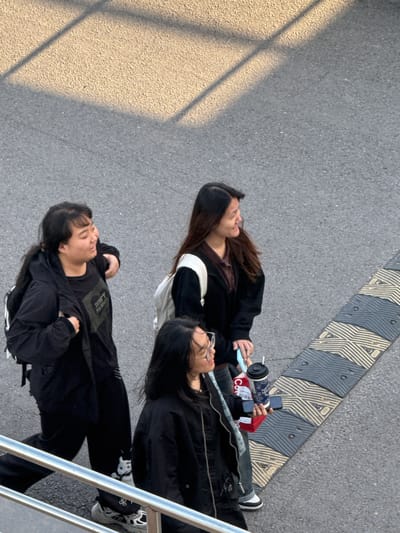 Three young adults are captured from an elevated perspective, resting on a paved outdoor surface in Zhongtun, China. The scene is bathed in direct sunlight, creating sharp, angular shadows across the grey asphalt ground, suggesting it is daytime with clear weather.

The individuals are lying or leaning back on the ground, facing upwards. On the upper left, a person with long dark hair, wearing a black jacket over a dark top, holds a light blue smartphone. Next to them, another individual wears glasses, a black jacket, and dark pants. They hold a blue-patterned reusable cup with a straw in one hand and a black smartphone in the other. This smartphone rests on a red object, possibly a small bag, which has the text "Co" visible on it. The third person, positioned lower in the frame, has dark hair and wears a black long-sleeved shirt with the white text "DON'T PANIC" printed across the chest.

The ground features a distinctive patterned strip of grey and yellow-striped segments, possibly a textured pedestrian crossing or a speed bump, running diagonally across the upper right of the frame. A metal railing is visible along the far right edge. The individuals appear calm and relaxed, taking a break in an urban or semi-urban setting.