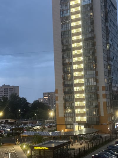 This image depicts a twilight scene in Kazan, Russia, featuring a tall residential building and a parking lot. The building, characterized by its beige and orange facade and numerous balconies with glass railings, is brightly illuminated from within, suggesting it is evening. In the foreground, a covered area with a modern, angled roof is visible, likely a communal space or shelter. Adjacent to this is a sign with Cyrillic text, "ВЕЛОПАРКОВКА," which translates to "BICYCLE PARKING." A large, paved parking lot filled with cars is situated behind this covered area, extending towards the base of the residential building. Further in the background, several other multi-story buildings and dense foliage are present, under a dusky, cloud-filled sky. The overall impression is of an urban residential environment in the evening.