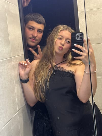 This is a horizontal, slightly distorted selfie taken in a mirror, showing two young people in Plovdiv, Bulgaria. The primary subject is a young woman with blonde hair, wearing a black dress with lace detailing. She is holding a smartphone in her right hand, taking the photo, and is winking at the camera with her lips pursed. Her left hand rests on her arm, and she is wearing a ring on her left ring finger and a bracelet on her left wrist.

Reflected next to her is a young man with short, dark hair and a mustache. He is lying down, with his head resting on a dark surface, possibly the frame of the mirror or a piece of furniture. He is making a peace sign with his right hand. His expression is relaxed and he appears to be looking towards the camera.

The setting appears to be indoors, with tiled flooring visible beneath them. The reflection suggests they are in a well-lit environment, possibly a bathroom or dressing room, given the tiled wall in the background. The overall mood is casual and playful. There is no visible text in the image.