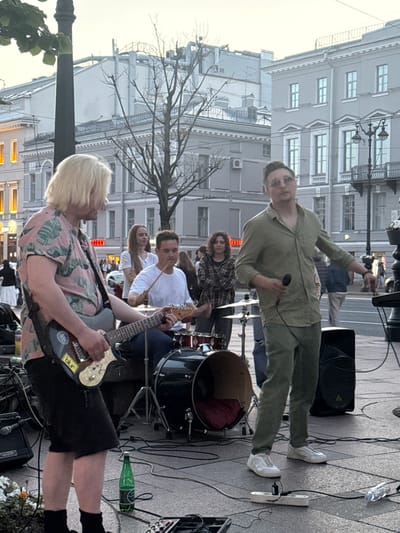 A band is performing live on a street in Saint Petersburg, Russia. The band members are playing guitars and drums. The lead singer is wearing a green shirt and pants and holding a microphone. A guitarist in the foreground has blond hair and is wearing a pink shirt with a leaf pattern. The scene appears to be set up on a paved area, possibly a sidewalk or square, with buildings in the background. The lighting suggests it is late afternoon or early evening, with a soft, diffused light. The mood seems to be casual and energetic, typical of a street performance. A green bottle of what appears to be beer is on the ground near some electrical equipment. There is some visible text on a sticker on the guitar that says "blur".