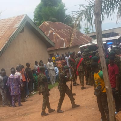 The image shows a scene in Akwanga, Nigeria. A crowd of civilians is gathered outside a building with a corrugated metal roof. Several uniformed soldiers with rifles stand among the civilians. Some soldiers have red sashes across their chests. An ambulance with blue lights is visible in the background. The ground is a reddish dirt. A tree trunk is visible in the foreground, partially obscuring the view.