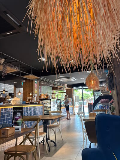 A woman is standing at a counter in a cafe. She is wearing a gray t-shirt and black shorts, and she is looking down at something on the counter. The cafe is decorated with natural materials, such as wood and straw. There are tables and chairs arranged throughout the space, and there are plants on shelves and hanging from the ceiling. The lighting is warm and inviting, and the overall atmosphere is relaxed and casual. Sunlight streams in through large windows, suggesting it is daytime. Some visible text includes "Cafe del Mar" and what appears to be product labels. The scene appears to be a modern and stylish cafe, possibly a popular spot for locals and tourists alike, given the European setting of Valencia, Spain.