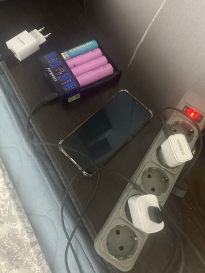 The image shows a close-up view of a power strip with multiple electrical outlets, a Samsung charging brick, and a Borofone charging brick. A smartphone is connected to the power strip, suggesting it is being charged. To the left of the phone, a battery charger with several batteries, including two pink and one blue, is also plugged into the power strip. The blue battery is labeled "2600mAh". The battery charger itself has a display screen that shows illuminated blue indicators. The power strip has a red on/off switch. The scene appears to be indoors, possibly on a desk or nightstand, with a light-colored textured surface visible in the background. The lighting suggests it could be daytime or evening, but there are no strong indicators of time of day. The overall impression is one of everyday electronic device charging activity. Visible text includes "SAMSUNG", "BOROFONE", and "2600mAh" on a battery, and "LiitoKala" on the battery charger. The location context is Odesa, Ukraine.