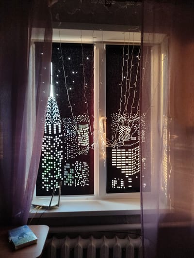 This media file depicts a window with a nighttime cityscape scene created on the blinds. The blinds are dark with cutouts that allow light to shine through, forming the shapes of skyscrapers and city lights. In the foreground, sheer purple curtains hang on either side of the window, partially obscuring the view. String lights are also draped across the window, adding to the nighttime ambiance.

On the windowsill, a white router with two antennas is visible, along with some cables connected to it. To the left of the window, on a small wooden table, rests a book with a blue cover, featuring an image of a person on what appears to be a horse. Below the window, a radiator is visible.

The overall scene conveys a sense of a cozy indoor setting at night, with a decorative, artificial cityscape providing a backdrop. The lighting, with the string lights and illuminated city scene, creates a warm and somewhat magical atmosphere. There is no visible text in the media file. The location context provided is Abinsk, Russia.