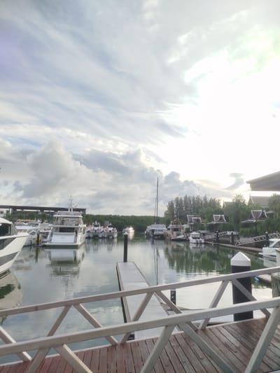 The image shows a marina in Ko Kaeo, Thailand, with several yachts and smaller boats docked. The sky is cloudy with patches of sunlight. In the foreground, a wooden pier with a white railing is visible. The water in the marina is calm and reflects the sky and the boats. In the background, there are trees and buildings with distinctive Thai architecture. No people are visible in the image. The overall scene depicts a peaceful marina setting.