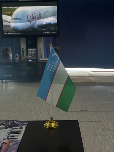 A small Uzbek flag on a gold stand is positioned in the foreground, angled towards the right. It is placed on a dark surface, possibly a table, where a magazine or booklet with images of people in uniform is partially visible on the left. In the background, a television screen displays an image of a Qatar Airways airplane. Below the television, a dark cabinet is visible with speakers on either side. A small statue and a framed photograph are placed on top of the cabinet. The overall setting appears to be an indoor space with tiled flooring. The scene is devoid of any people or discernible activity.