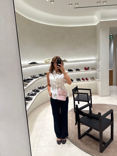 A woman is taking a mirror selfie in a modern, well-lit shoe store, likely a boutique or department store. She is wearing a white sleeveless blouse, dark wide-leg pants, and black woven sandals. She is accessorizing with a pink quilted crossbody bag with a gold chain strap and a gold bracelet. She has shoulder-length brown hair and is wearing glasses.
The store environment features sleek, curved shelves displaying various shoes, primarily sandals and flats. The walls are a light, textured neutral color, and the ceiling has recessed lighting. In the foreground, two minimalist black chairs with leather straps are placed on a circular rug. The floor is tiled with large, light-colored square tiles. A large mirror on the left reflects the scene. The overall aesthetic is clean, minimalist, and upscale. The image does not provide any specific location details within Loures, Portugal.