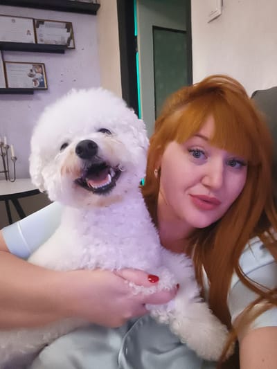 The image shows a woman with long, reddish-orange hair and bangs, and a white bichon frise dog. They are both looking at the camera. The woman is wearing a light blue top. Her makeup includes pink lipstick and eyeliner. She has a slight smile and her eyes are open. The dog is smiling with its mouth open and tongue visible. The woman is holding the dog, and its paws are resting on her shoulder and arm.
In the background, there are shelves on the wall with framed certificates. The lighting suggests it might be indoors, possibly in a home or office setting. The overall mood appears to be casual and affectionate, with the woman and her dog posing together for a picture. There is no visible information about the time of day or weather. The location context is Odesa, Ukraine, but there are no specific visual cues in the image to confirm this.