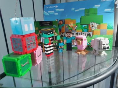 The image displays a collection of Minecraft-themed toys and merchandise arranged on a glass table, possibly in a home setting. The subjects are primarily Minecraft characters and creatures, rendered in their characteristic blocky style. These include figures of Steve, a pink sheep, a white wolf, and a Creeper. There are also stacking toy blocks in green, red, and blue, some of which appear to be containers for small toys. A key item in the foreground is a green toy block with an image of a pickaxe on its side.

In the background, a large blue board with Minecraft imagery and the "Carrefour" logo suggests a promotional display or packaging, possibly from a collection or album. The overall scene is bright and colorful, with toys scattered casually. The lighting suggests it is daytime. There is no discernible weather information, but the indoor setting is evident. The presence of the Carrefour logo could indicate a retail or promotional context, perhaps in Romania given the provided location context.

Visible text includes "Carrefour", "A MINECRAFT MOVIE", and "MINECRAFT" on the various items. The toys are static, indicating that no specific action is taking place beyond their arrangement. The overall impression is one of playful display of popular video game merchandise.