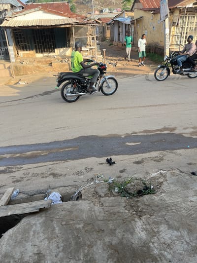 In an urban setting in Abuja, Nigeria, an elderly man wearing a green t-shirt and a grey baseball cap is riding a motorcycle. The motorcycle is black with chrome accents and has a license plate visible. The man is looking ahead as he rides. Behind him, two people are walking. One is a man wearing a green t-shirt and dark pants, while the other is a woman wearing a white shirt and a green skirt, holding something in her hand. Further in the background, another motorcycle is parked, with two people on it. The street is paved and there are buildings on either side, with corrugated iron roofs. Some of the buildings appear to be shops. The area shows signs of wear and tear, with debris on the street and some of the buildings in disrepair.