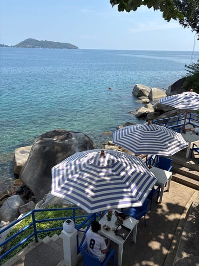 The image shows a seaside dining area in Kamala, Thailand. Three blue and white striped umbrellas provide shade over tables and chairs placed alongside the rocky shoreline. The tables are set with dining items. One person wearing a white shirt with the number 8 is seated at a table. The calm, clear turquoise water of the Andaman Sea stretches out towards the horizon, with a distant landmass visible. Large, smooth rocks are interspersed among the tables and chairs, partially submerged in the water. A set of blue railing along the right of the image indicates a stair way along the edge. Above the railing are trees that cast a shadow.