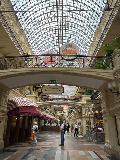 The image shows an interior view of a large, ornate building with a glass ceiling. The scene appears to be a shopping arcade or department store, likely in Moscow, Russia, given the visible Russian text.
Several people are walking through the arcade. In the foreground, a man in a white t-shirt, blue jeans, and a white baseball cap walks away from the camera, carrying an orange bag. Another person, wearing a white shirt and dark pants, walks towards the camera further down the arcade. Other individuals are visible in the background, some appearing to be shoppers and others possibly staff.
The architecture is grand, with arched walkways, decorative moldings, and ornate lamps. The curved glass ceiling allows natural light to illuminate the space. There are various shops or stalls lining the arcade, some with awnings. One awning is striped in red and white.
Visible text includes signs in Russian. One sign with an arrow reads "к Фонтану" which translates to "to the Fountain," with an English translation "TO THE FOUNTAIN" below it. Another sign shows "ЛУКОВЫЕ ЛИНИИ" which translates to "Onion Lines." There is also a circular sign with an image of ice cream cones and Russian text.
The time of day appears to be daytime, as suggested by the natural light. The weather outside is not discernible, but the interior is well-lit. The overall impression is one of a busy, elegant public space.