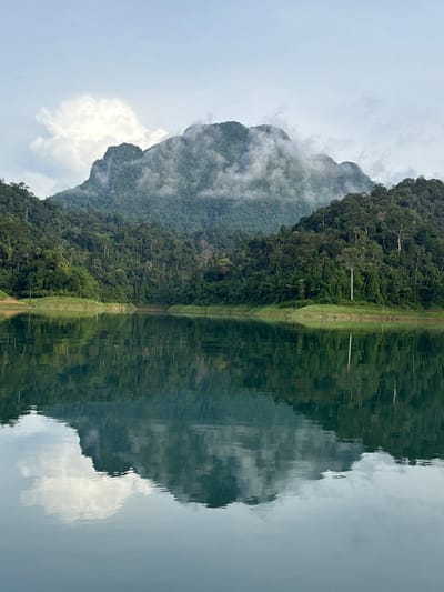 The image depicts a serene natural landscape at Khao Phang, Thailand, characterized by a body of calm water reflecting lush green mountains and a cloudy sky. The foreground is dominated by the still water, which perfectly mirrors the scene above. In the middle ground, the tree-covered slopes of the mountains rise from the water's edge, creating a dense, verdant wall. The mountains themselves are rugged, with their peaks partially obscured by wisps of white cloud. The sky above is overcast, with soft grey clouds interspersed with patches of lighter sky. The overall impression is one of tranquility and natural beauty, typical of the landscape found in this region of Thailand. There are no people or man-made structures visible in the immediate scene, emphasizing the untouched quality of the environment.