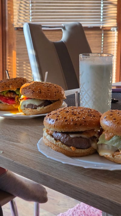 The image shows two burgers on a wooden surface, along with a glass of what appears to be a milky drink. The burgers are sesame seed buns, and one of them has a toothpick inserted. The scene is indoors, likely a restaurant or a home setting, with blinds visible in the background. The lighting suggests it could be daytime. A person's legs, wearing socks, are visible in the lower right corner of the frame, suggesting they are seated at the table. The overall mood is casual and relaxed, with the focus on the food and the implied dining experience. No text is visible in the image.