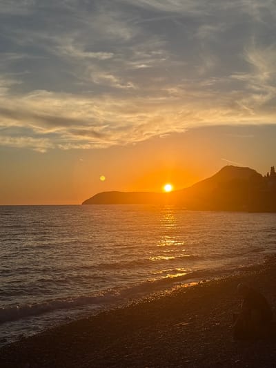 The image depicts a serene sunset scene at a beach in Bar, Montenegro. The sun, a brilliant orange orb, is setting behind a silhouetted hill, casting a warm golden glow across the sky and the water. Wispy clouds are scattered across the upper half of the frame, tinged with the colors of the sunset. The sea is calm, with gentle waves lapping at the pebble shore. In the foreground, on the right side of the image, a lone figure is seated on the beach, facing away from the camera and towards the sea, appearing to be contemplating the sunset. The beach is covered in dark pebbles. The overall atmosphere is peaceful and tranquil.