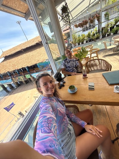 The image depicts a couple seated at a wooden table in an open-air restaurant overlooking a beach and ocean in Ko Tao Subdistrict, Thailand. In the foreground, a woman with curly brown hair smiles at the camera, wearing a long-sleeved, multicolored top and denim shorts. To her right, a man with short brown hair and a patterned t-shirt is seated, looking towards the camera.

On the table, a green drink in a cup, a purple drink, a smartphone, and a menu are visible. In the background, through a large glass window, a thatched-roof building with outdoor seating and a sandy beach can be seen. The restaurant appears to be decorated with plants hanging from the ceiling and shelves. The lighting suggests it is daytime.