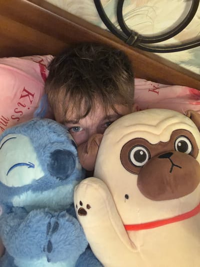 In a bedroom, a person, likely male, peeks out from behind two plush toys: a blue Stitch character on the left and a light brown pug on the right. The person's eyes are visible, and their hair is dark and messy. The plush toys are large and occupy most of the frame. The background includes a wooden headboard and a pink pillow with red lettering that appears to say "Kiss me." The overall impression is of a cozy, personal space. This image was not taken in Saint Petersburg, Russia.