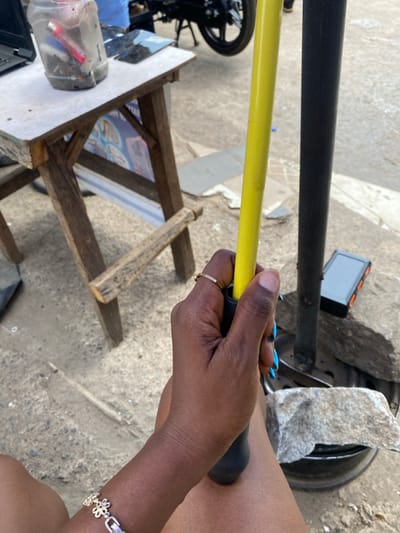 The image depicts an outdoor street scene in Ibadan, Nigeria, characterized by a mix of commercial activity and utilitarian elements. In the immediate foreground, a dark-skinned human hand grasps a yellow pole with a black rubber grip. A thin gold-colored ring is present on the ring finger, and fingernails are painted light blue. A silver-colored bracelet with interconnected flower-shaped links adorns the wrist. The person's bare upper thigh is visible below the hand, suggesting a seated or resting posture.
To the left of the frame, a weathered wooden table with a white laminated top serves as a makeshift workspace. On the table, a clear plastic jar, likely a repurposed bottle, contains miscellaneous items including a red flashlight and other small objects, possibly tools or debris. A black laptop is partially visible on the far left of the table, and several pieces of cracked or disassembled glass, resembling phone screens, are laid flat. Behind the table, a partially visible banner or poster displays graphics that appear to be electronic components.
Further into the scene, a black vertical pole stands to the right of the yellow pole, possibly another support structure. Near its base, a compact black and orange electronic device, possibly a power bank or portable modem, rests on the ground. A gray rock sits atop what appears to be a discarded black tire rim, also on the ground. The ground surface consists of rough, light-colored dirt or concrete. In the background, a motorcycle tire and a portion of a black motorcycle are visible on the right, and a person's legs wearing light blue pants are partially visible behind the table. The overall lighting is bright, indicative of daylight.