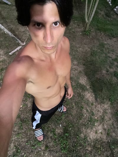 A shirtless man with dark hair stands outdoors, looking directly at the camera. He is wearing black shorts with white stripes and striped sandals. The man's body is tanned, with visible abdominal muscles and a tattoo on his left forearm. He is positioned on a grassy and earthy ground, with some scattered twigs and leaves. In the background, a thin tree with sparse foliage is visible, along with some out-of-focus vegetation and what appears to be a fallen branch or piece of wood. The lighting suggests it is either dusk or dawn. The specific location within Los Velásquez, Venezuela cannot be determined from the visual information alone.