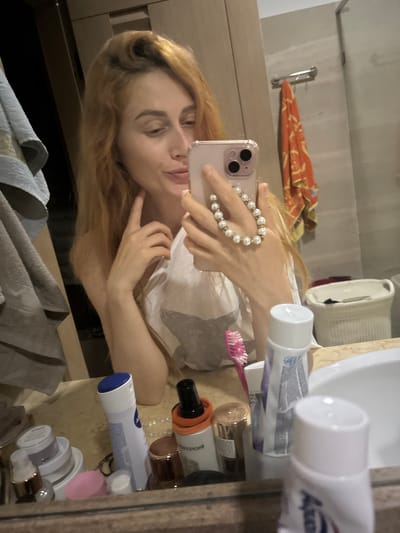 A young woman with long, reddish-blonde hair is taking a selfie in a bathroom mirror. She is wearing a white t-shirt and has her left hand raised to her chin, while holding a pink smartphone in her right hand. A pearl bracelet adorns her right wrist. The bathroom appears to be well-lit and stocked with various toiletries, including bottles of Nivea lotion and other cosmetic products on the counter. A pink toothbrush is also visible. In the background, there are towels hanging and an orange towel draped over a rack. The overall scene suggests a moment of personal grooming or preparation.
