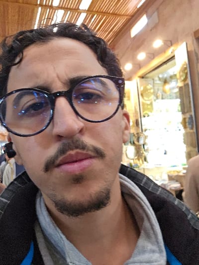 The image is a vertical close-up selfie featuring an adult male from the chest up. He has dark, curly hair, a mustache, and a goatee. He wears dark-rimmed circular glasses, which show prominent blue light reflections. His attire includes a grey polo shirt beneath a dark jacket with a visible patterned collar. His brows are slightly furrowed. The background identifies an indoor market or souk environment, consistent with locations in Marrakesh, Morocco. Overhead, a slatted wooden ceiling is visible. To the subject's right, a brightly lit shop interior displays various items, possibly metalware or ceramics, illuminated by spotlights. A second, partially obscured individual is visible in the distant left background.