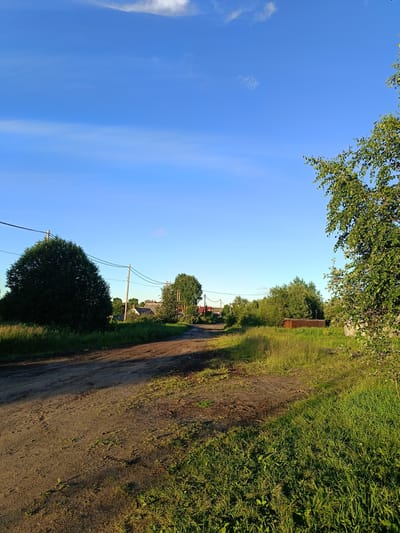 The image depicts a rural road in Arkhangelsk, Russia, under a clear blue sky with sparse, wispy clouds. The road is unpaved and appears to be made of dirt and gravel, with visible tire tracks and some patches of exposed earth. Lush green grass and vegetation line both sides of the road, with shadows from trees cast across the ground.
To the left of the road, a large, dense, dark green tree dominates the foreground. Further down the road, several houses with visible roofs are scattered among more trees and greenery. Utility poles with electrical wires are present along the left side of the road, contributing to the rural aesthetic. On the right side of the road, another tree with lighter green foliage is visible, and a brown, rectangular structure, possibly a shed or storage container, is situated in the grassy area. The overall scene suggests a quiet, natural environment on the outskirts of a settlement. No people or animals are visible in the image.