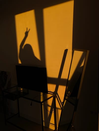 The media file depicts a shadow of a person making a peace sign on a wall bathed in warm, golden light. The light appears to be sunlight, suggesting it's either sunrise or sunset. The wall is lit by a rectangular source of light, possibly a window. The shadow of a hand with two fingers extended in a "V" shape is clearly visible. To the right, a dark object, possibly a monitor or a picture frame, casts its own shadow. Above, there's a reflection in what appears to be a mirror or a glass surface, with a metallic frame. The overall scene is dimly lit, with the golden light creating a dramatic contrast with the darker areas. The setting is likely indoors, given the presence of furniture-like structures and a monitor. The mood evoked is calm and contemplative, with the warm light and the peace sign suggesting a moment of peace or reflection. No text is visible in the image. The location context of Izhevsk, Russia, is provided but not visually discernible within the image itself.