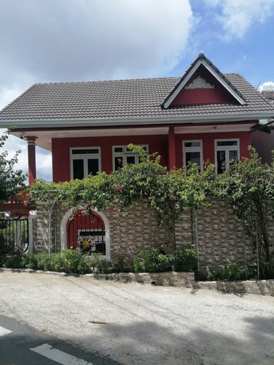 The image shows a red two-story house with a gray tiled roof in the city of Da Lat, Vietnam. The house has white-framed windows and a prominent gable with decorative molding. A stone wall with a decorative arched entrance separates the house from the street. Lush green foliage, including flowering vines, grows over the wall and around the property. A black metal gate is visible on the left side of the wall. The ground in front of the house is paved with concrete and appears to be a sloped driveway or street. The sky is partly cloudy with blue patches. No people are visible in the image.