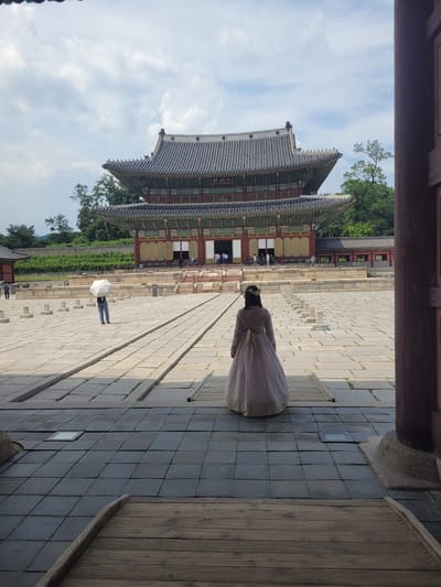 The image shows a view of Gyeongbokgung Palace in Seoul, South Korea. In the foreground, a woman wearing a traditional Hanbok dress stands facing away from the camera, looking towards the palace. The palace is a large, traditional Korean building with multiple tiered roofs and intricate architectural details. It is situated in a large, open courtyard paved with stone. Several other people are visible in the background, some walking and others observing the palace. One person in the middle ground holds a white umbrella. The sky is partly cloudy. The scene captures a moment of cultural tourism at a historical landmark.