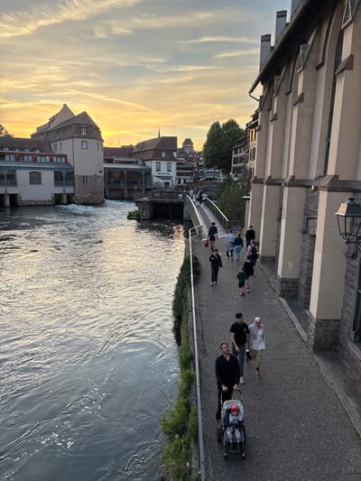 This image depicts the Ponts Couverts and the Barrage Vauban in Strasbourg, France, during sunset. The sky is filled with warm orange and yellow hues from the setting sun, with scattered white clouds and contrails. The Ill river flows through the foreground, its surface rippled and reflecting the colors of the sky.

On the right side of the image, a light-colored building with a textured facade and arched windows stands prominently. A pedestrian walkway runs alongside the river, bordered by a low wall and greenery. Several people are walking on this path.

In the middle ground, the Ponts Couverts are visible, a series of three medieval stone bridges with three towers. The Barrage Vauban, a large dam with archways, is situated further upstream, with water flowing through its structure. Several buildings with traditional Alsatian architecture, characterized by half-timbered facades and steep roofs, line the riverbanks in the background.

A diverse group of people are captured in motion on the walkway. In the foreground, a man is pushing a child in a stroller, followed by a couple. Further up the path, other individuals and groups are walking in various directions, suggesting a leisurely stroll or commute. The overall scene conveys a sense of tranquility and urban charm at dusk.
