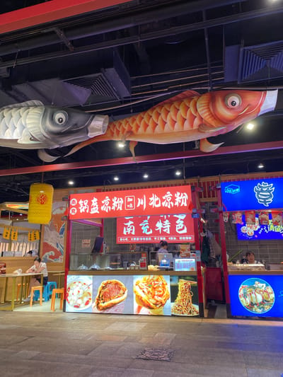 In Qingyang District, China, an outdoor food stall area is depicted. The scene is dominated by a large red and white sign above a food stall, featuring Chinese characters and images of food. Two large, inflatable koi fish decorations are suspended from the ceiling, one grey and one orange, both wearing face masks. The food stall itself displays illuminated panels showcasing various dishes, including what appears to be a bowl of soup with dumplings, a large sandwich or bun filled with meat, a dish with shrimp, and a plate of noodles. In the background, other food stalls with blue signage are visible. To the left, a woman sits at a small table, facing away from the camera, while a vendor stands behind the counter of a stall to the right. The ground is paved with stone tiles.