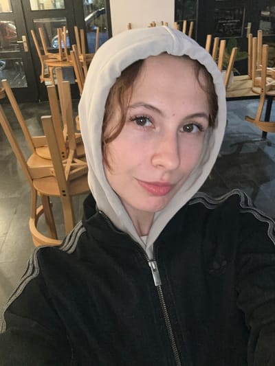 A selfie of a young woman in a white hooded sweatshirt and a black jacket with light gray stripes on the sleeves. She is looking at the camera with a slight smile. The background appears to be a restaurant or cafe, with stacked wooden chairs. Based on the style of the chairs and the general ambiance, it is potentially a restaurant or cafe located in Novi Sad, Serbia.