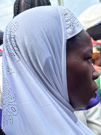 A close-up shows a person of African descent wearing a white head covering, possibly a hijab, adorned with small, metallic studs or rhinestones. The person's face is visible from the nose up. The background is somewhat blurred, but it seems to depict other people and suggests the setting might be outdoors, possibly in Lekki, Nigeria.