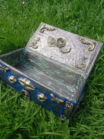An open, ornate, rectangular box sits on bright green grass. The box has a dark, textured interior with streaks of teal and black. The exterior is primarily dark blue with metallic gold decorative elements. These elements include what appear to be seashells, small winged insects, and floral motifs, such as roses. The lid of the box is open and also features metallic gold decorative elements, including scrollwork and floral designs. The box is positioned outdoors on a grassy area, suggesting it is in a garden or park. The background consists of more dense green grass. The scene is devoid of any human presence or activity. The lighting suggests it is daytime.