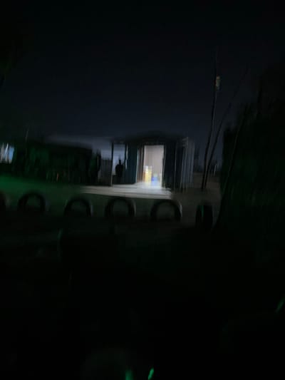 The image depicts a dimly lit outdoor scene at night, likely in a rural or semi-urban area. In the background, a small, open-fronted structure, possibly a shed or makeshift stall, is visible. The interior is illuminated, revealing shelves with what appear to be bottles or containers. A silhouetted figure stands just inside the entrance. In the foreground, a row of what seem to be repurposed tires, cut in half and placed upright, forms a barrier or decorative element. The overall impression is one of a slightly disheveled, possibly abandoned or under-construction site, with sparse lighting adding to a somewhat eerie atmosphere. The location is identified as Howayi, Nigeria.