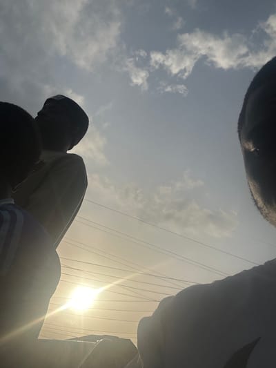 The low-angle shot captures the faces of a group of people standing in Umuode, Nigeria. The sun is shining brightly, positioned near power lines. Clouds are visible in the sky.