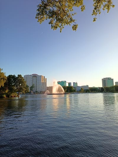 A wide, calm body of water fills the foreground and mid-ground of the image. The water has a dark blue hue and is rippled by a gentle breeze, reflecting the clear blue sky and the surrounding environment. In the mid-ground, a large fountain sprays a dramatic plume of water upwards, illuminated by the warm, golden light of what appears to be late afternoon or early morning sun. Beyond the fountain, a city skyline is visible. Dominating the left side of the skyline is a tall, multi-story white building with numerous windows. To the right of this building, other modern structures, including those with green glass facades, are present. Trees line the edge of the water on the left side, with their foliage catching the sunlight. Overhead, branches of a leafy tree hang into the frame from the top, casting dappled shadows. The sky is a clear, pale blue, with a faint crescent moon visible near the top left. The scene suggests a park or waterfront area in a city, likely a public space for recreation and leisure. This scene depicts the Zaslavskoye Reservoir, also known as Minsk Sea, in Minsk, Belarus.
