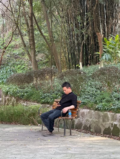 The image depicts an outdoor scene in Jiangjin, China. A male individual, appearing to be of East Asian descent, is seated on a slatted wooden park bench. He is dressed in a black long-sleeved turtleneck shirt, dark blue jeans, and gray athletic shoes. The man holds a dark mobile phone in his right hand, his head tilted downward as if viewing the device. A clear plastic water bottle with a blue cap rests on the bench to his left. The bench is situated on a gray paved surface. Behind the bench, a stone retaining wall supports a rise in terrain covered with dense green foliage, including shrubs and small trees. Further into the background, a hillside covered with diverse trees and a prominent stand of tall bamboo stalks is visible. The lighting is diffused, suggesting an overcast day or shaded area. No other individuals are present in the frame.