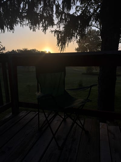 A folding chair is positioned on a wooden deck facing a sunrise over a grassy field. The scene is silhouetted against the bright sky, with tree branches hanging overhead. The deck appears to be part of a residential property in Holt, Michigan, United States. The chair is green and made of fabric and metal. The deck is made of weathered wooden planks. The grassy field has some scattered trees and a fence in the distance. The time of day is early morning, indicated by the low angle of the sun.
