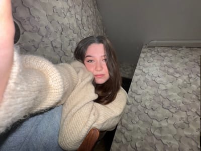 A young Caucasian woman with long, dark brown hair is taking a selfie indoors. She is wearing a beige, knitted sweater and blue jeans. Her head is tilted, and she is resting her cheek on her arm. Her eyes are open and looking at the camera. The background appears to be a room with floral patterned wallpaper. The lighting is dim. The image is a close-up shot, with the woman's face and upper body filling most of the frame. The location is specified as Sakmary, Russia.