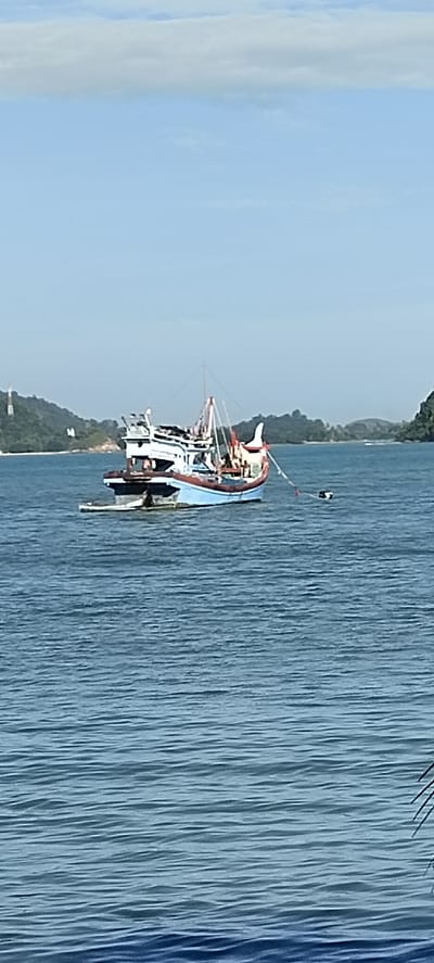A medium-sized fishing boat with a blue hull and white superstructure is anchored in the sea off the coast of Lhoktimon, Indonesia. The boat has a mast with rigging and several people can be seen on deck. In the background, a hilly, tree-covered coastline is visible, with a clear blue sky above. The water is calm with gentle ripples. A small black and white buoy is attached to a rope from the boat.