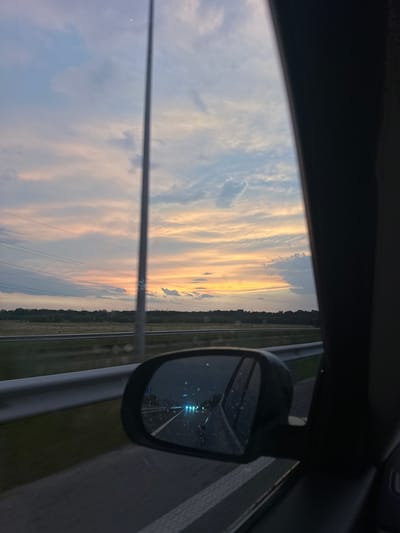 The image captures a view from a car window in Kvitneve, Ukraine, during sunset. The sky is a canvas of pastel colors, with streaks of pink, orange, and blue clouds illuminated by the setting sun. A tall, thin pole stands vertically in the center of the frame, piercing the sky. Power lines crisscross the horizon, against a backdrop of a dark, wooded landscape. In the foreground, a car's side mirror reflects the road behind, showing the lights of oncoming vehicles and what appear to be water droplets on the mirror's surface. The overall impression is one of a serene, dusky journey on the outskirts of Kvitneve.