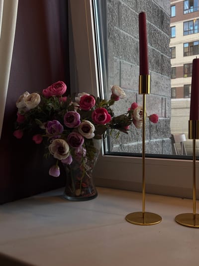 A vase of artificial flowers, predominantly pink and white, sits on a windowsill. Two red tapered candles in gold holders are positioned to the right of the vase. Through the window, a textured grey building with multiple windows is visible, suggesting an urban environment. The scene is likely indoors, with a curtain on the left side of the frame. There is no indication of people or any specific location within Saint Petersburg, Russia.