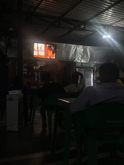A dimly lit outdoor bar in Jos, Nigeria. Two people are sitting on green plastic chairs with Heineken logos. One person is wearing a checkered shirt. Another person is sitting at the bar near a Heineken branded refrigerator. A television on the wall displays a brightly lit image. The bar has a corrugated metal roof. There is a bright light under the roof. There are various objects and supplies near the bar area, including cardboard boxes and equipment under a table. The atmosphere appears casual.