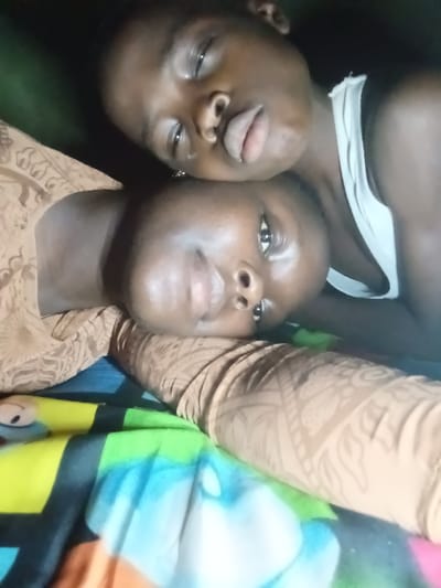 Two young, dark-skinned people are close together in a dimly lit setting. One wears a white tank top and seems to be leaning or resting on the other. The second person is wearing a patterned brown garment, possibly a shirt or dress. They are lying on colorful bedsheets. Both subjects are looking toward the camera. The focus appears to be on the person with their head tilted.