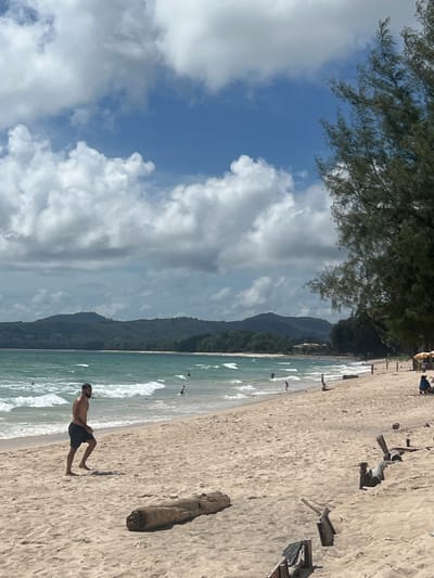 A man is jogging on a sandy beach in Choeng Thale, Thailand. The sky is partly cloudy with bright blue patches. The ocean has green waves, and there are several people in the water and on the beach. A large tree with green foliage is on the right side of the image, and there are some pieces of driftwood and debris on the sand. In the background, there are lush green hills and mountains. The scene appears to be a sunny day at a tropical beach.