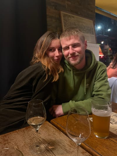 A man and a woman pose for a photo at a restaurant. The man wears a green hoodie and has a short beard. The woman has long brown hair and wears a black shirt. On the wooden table are wine glasses and a beer glass. Another person is partially visible in the background. The photo is taken indoors, with a window and some wall decorations visible.