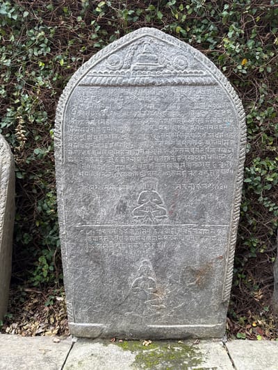 The image shows a gray stone slab with inscriptions in a non-English script. The slab is roughly rectangular with a rounded top edge and is set in a surrounding of greenery. The surface of the slab is aged and weathered. The slab is located outside in a natural setting, among plants. The location is Lalitpur, Nepal.